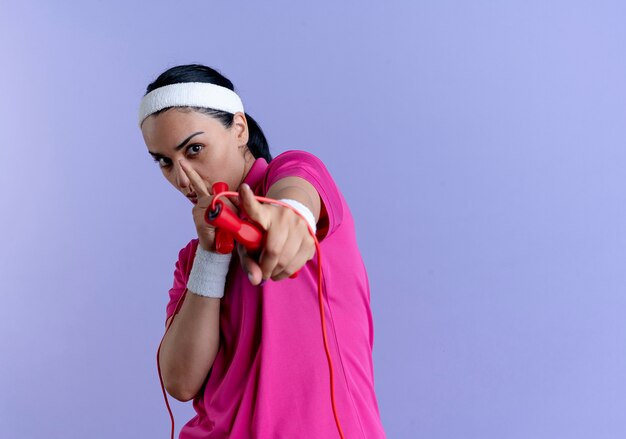 Молодая уверенная в себе кавказская спортивная женщина с повязкой на голову и браслетами держит скакалку и указывает на камеру