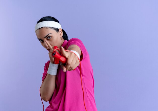 Young confident caucasian sporty woman wearing headband and wristbands holds jumping rope and points at camera