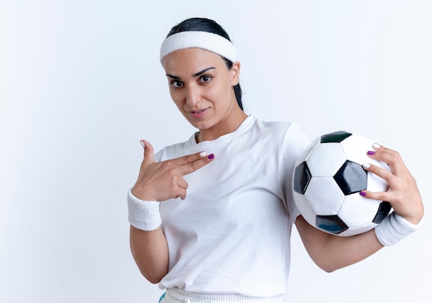 Young confident caucasian sporty woman wearing headband and wristbands holds and points at ball isolated on white space with copy space