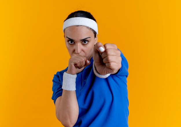 Young confident caucasian sporty woman wearing headband and wristbands keeps fists pretending to punch on orange  with copy space