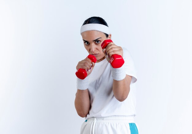 Young confident caucasian sporty woman wearing headband and wristbands pretends to defend holding dumbbells isolated on white space with copy space