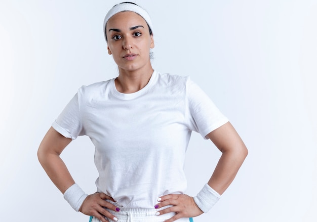 Young confident caucasian sporty woman wearing headband and wristbands puts hands on waist isolated on white space with copy space