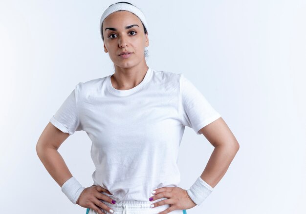 Молодая уверенная в себе кавказская спортивная женщина с повязкой на голову и браслетами кладет руки на талию, изолированную на белом пространстве с копией пространства