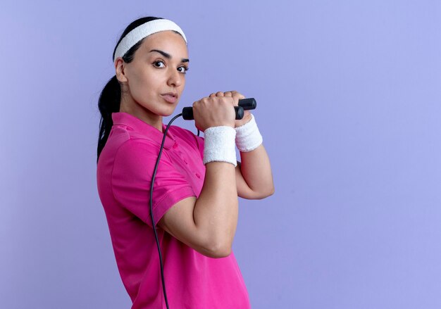 Young confident caucasian sporty woman wearing headband and wristbands stands sideways holding jumping rope on purple  with copy space