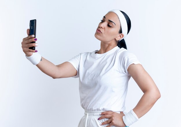 Молодая уверенная в себе кавказская спортивная женщина с повязкой на голову и браслетами стоит боком, глядя на телефон, делает селфи, изолированное на белом пространстве с копией пространства