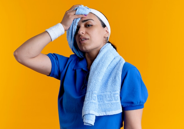 Young confident caucasian sporty woman wearing headband and wristbands with towel on neck wipes his forehead with a towel on orange with copy space