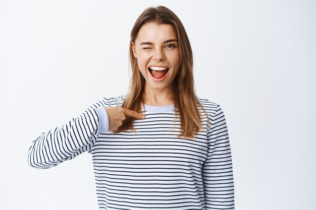 Young confident and cheeky woman winking at front, pointing at herself and smiling determined, self-promoting personal achievement and skills, white wall