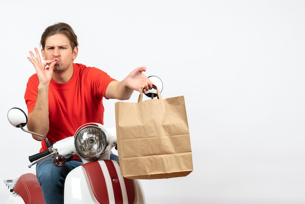 Young confident courier guy in red uniform sitting on scooter giving paper bag making perfect gesture on white wall