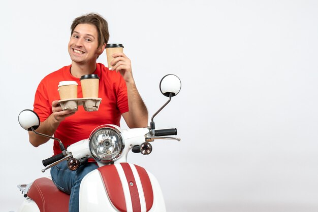 Young confident courier man in red uniform sitting on scooter and holding orders on yellow wall