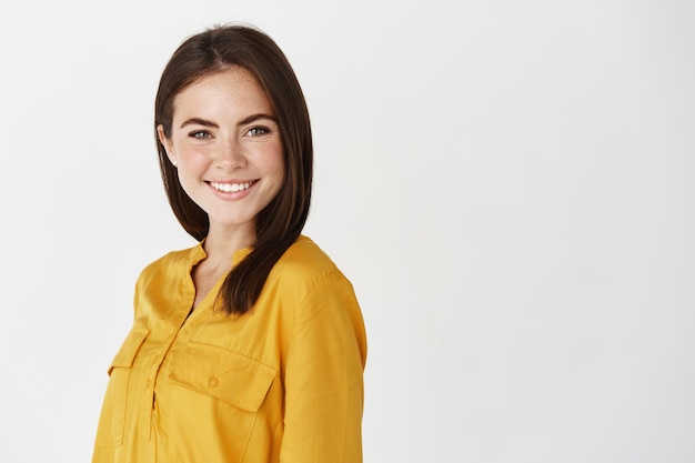 Young confident female manager smiling at front, standing near copy space for company logo, white wall.