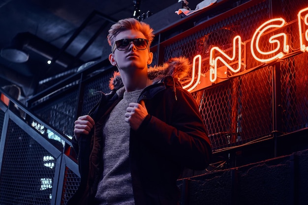 Young confident guy wearing coat with fur hood and sunglasses standing on the staircase to the underground nightclub, a backlit signboard in the background