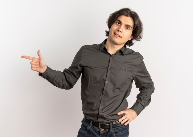 Young confident handsome caucasian man points at side and puts hand on waist isolated on white background with copy space