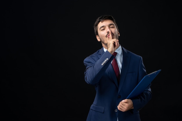 Young confident male office worker in suit holding documents and making silence gesture on isolated dark wall