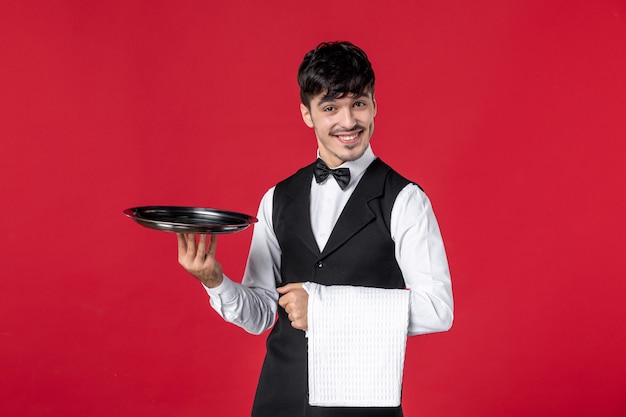 young confident male waiter in a uniform with butterfly on neck holding tray and towel on red background