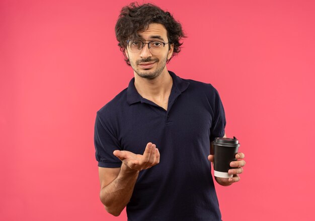 光学メガネと黒のシャツを着た若い自信のある男は、ピンクの壁で隔離のコーヒーカップとポイントを保持します。