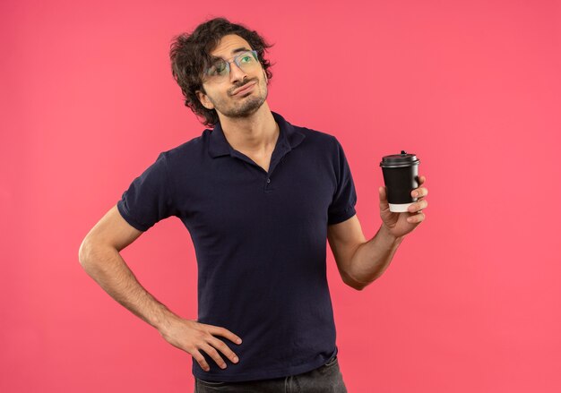 Young confident man in black shirt with optical glasses holds coffee cup and puts hand on waist isolated on pink wall