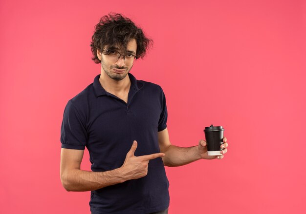 Young confident man in black shirt with optical glasses holds and points at coffee cup isolated on pink wall