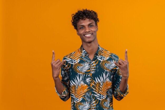 Young confident man holding hands in a gesture of rock music fan to express emotion of success on an orange background