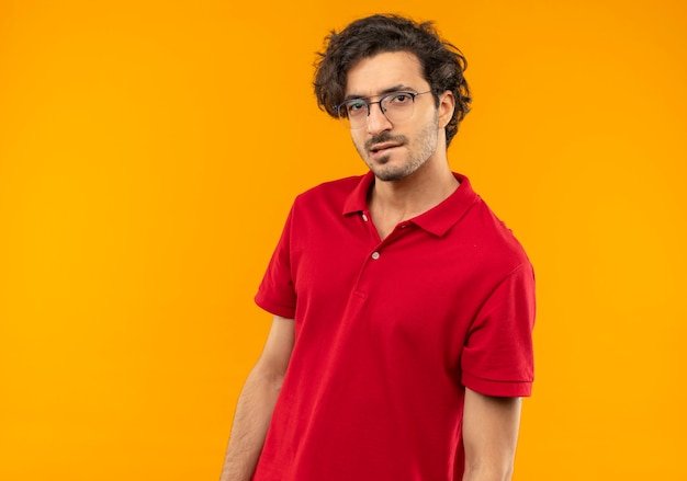 光学メガネと赤いシャツを着た若い自信のある男は唇を噛み、オレンジ色の壁に孤立して見える
