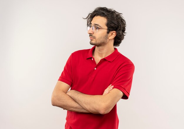 光学メガネと赤いシャツを着た若い自信のある男は腕を組んで白い壁に隔離された側を見る