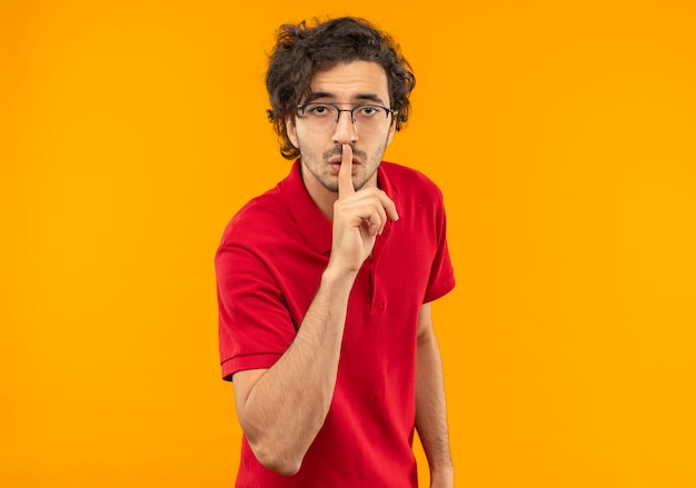 光学メガネのジェスチャーで赤いシャツを着た若い自信のある男は、オレンジ色の壁に孤立した沈黙のサインを維持します