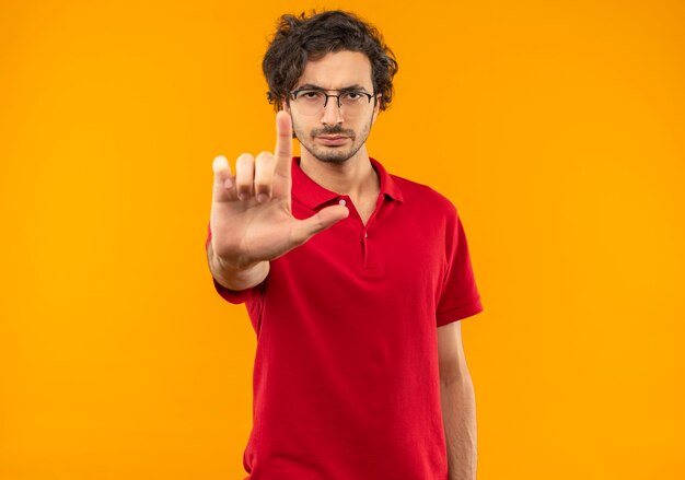 Young confident man in red shirt with optical glasses holds hand out and points up isolated on orange wall
