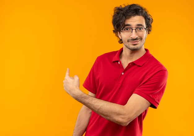 光学メガネと赤いシャツを着た若い自信のある男は後ろを指し、オレンジ色の壁に孤立して見える
