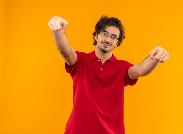 Young confident man in red shirt with optical glasses points with both hands isolated on orange wall