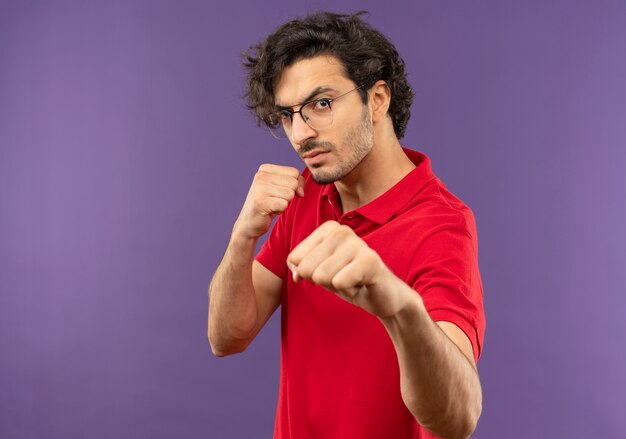 Young confident man in red shirt with optical glasses stands sideways and with fists up and ready to punch isolated on violet wall