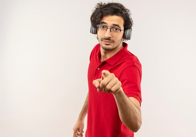 Young confident man in red shirt with optical glasses and with headphones points isolated on white wall