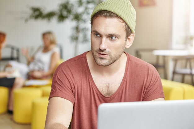 Молодой уверенный в себе мужчина сидит в кафе и работает на ноутбуке, задумчиво смотрит в сторону, пытаясь задействовать воображение