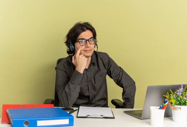 Young confident office worker man on headphones in optical glasses sits at desk with office tools using laptop points at eye