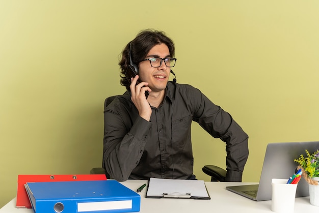 Foto gratuita uomo giovane lavoratore di ufficio fiducioso sulle cuffie in vetri ottici si siede alla scrivania con strumenti di ufficio utilizzando colloqui di laptop sul telefono