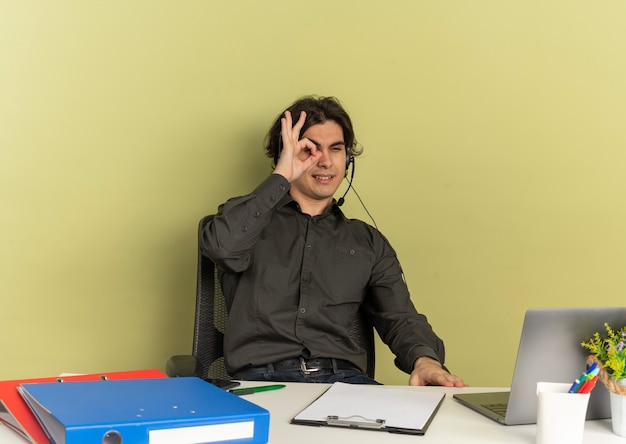 Молодой уверенный офисный работник человек в наушниках сидит за столом с офисными инструментами, используя и глядя сквозь пальцы на ноутбук