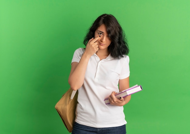 Young confident pretty caucasian schoolgirl wearing back bag points at eye with finger on green  with copy space