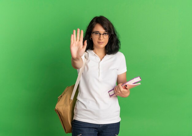 Young confident pretty caucasian schoolgirl wearing glasses and back bag gestures stop hand sign holding books on green  with copy space