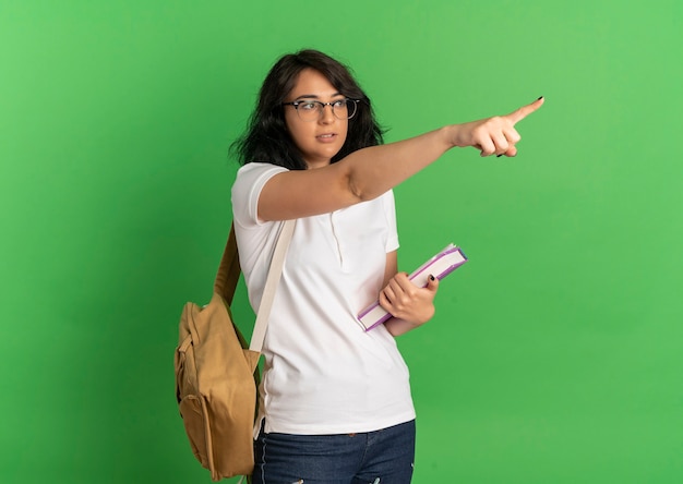 Young confident pretty caucasian schoolgirl wearing glasses and back bag looks and points at side holding books on green  with copy space