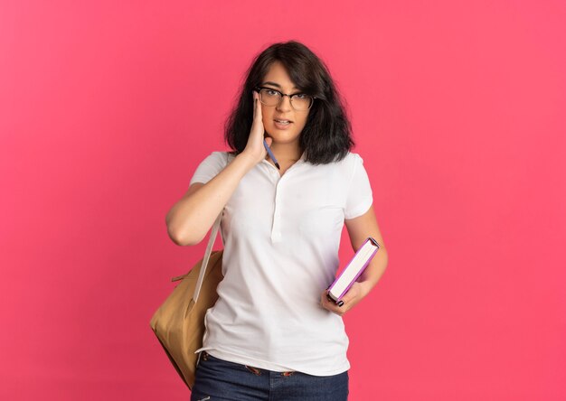 Young confident pretty caucasian schoolgirl wearing glasses and back bag puts hand on face holding pen and book on pink  with copy space