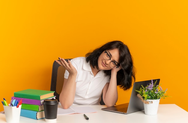 Young confident pretty caucasian schoolgirl wearing glasses sits at desk with school tools puts head on hand isolated on orange space with copy space