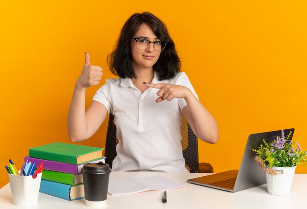 眼鏡をかけている若い自信を持ってかなり白人の女子高生は、学校の道具の親指を上にして机に座って、コピースペースでオレンジ色の側を指しています