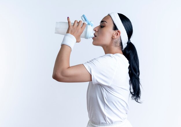 Young confident pretty sporty woman wearing headband and wristbands stands sideways drinking water isolated on white space with copy space