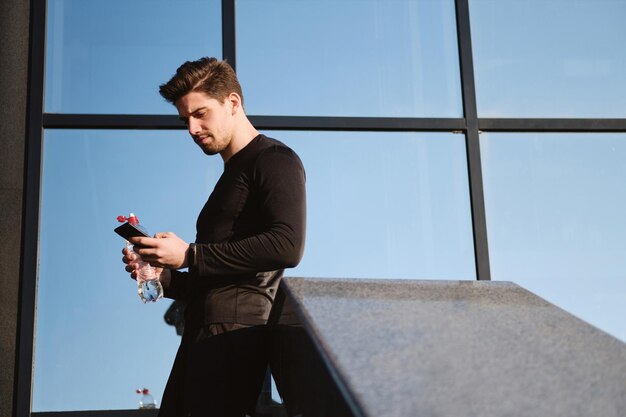야외 운동 후 휴대폰을 사용하여 순수한 물 한 병을 든 자신감 있는 젊은 스포티한 남자