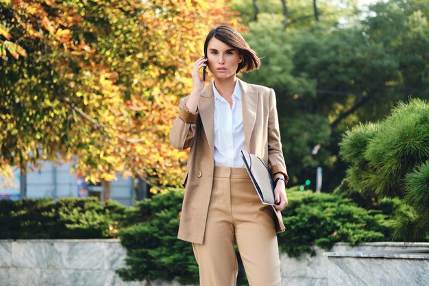 Young confident stylish businesswoman talking on cellphone intently looking in camera on city street