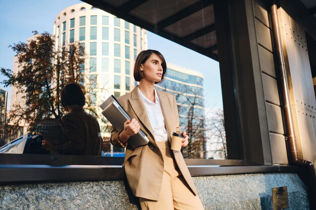 Young confident stylish businesswoman with laptop and coffee to go thoughtfully looking aside on city street