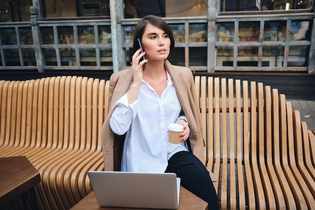 Young confident stylish businesswoman with laptop talking on cellphone during coffee break in cafe on street