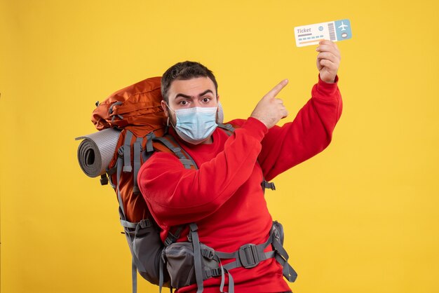 Young confident traveller guy wearing medical mask with backpack and holding ticket on yellow