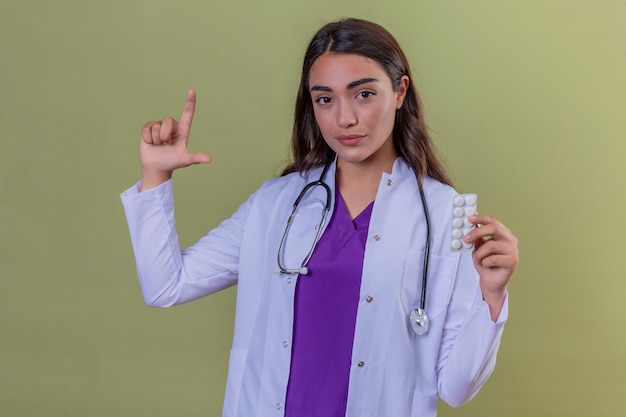 Young confident woman doctor in white coat with phonendoscope holding blister with pills and doing small size sign with fingers looking at the camera over green isolated background