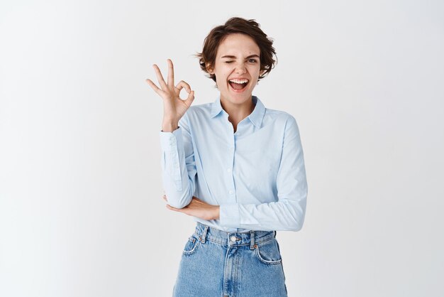 Young confident woman winking motivated and showing OK gesture approve and like product guarantee good quality standing on white background