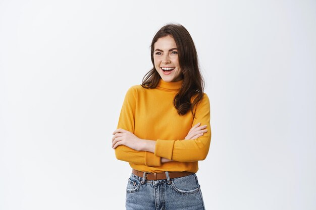 Young confident woman with natural light make up, cross arms on chest and staring aside at copy space, laughing happy on white