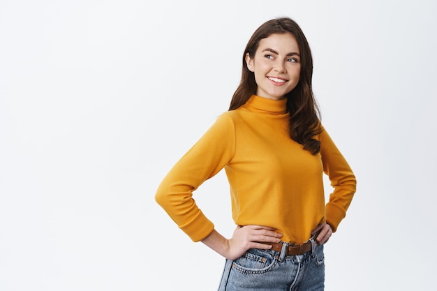 Young confident woman with natural light make up, holding hands on waist and looking left at empty copy space, staring at logo and smiling, standing on white wall